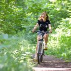 A woman cycling on a scenic forest trail surrounded by green foliage, enjoying a sunny day.