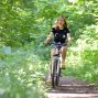 A woman cycling on a scenic forest trail surrounded by green foliage, enjoying a sunny day.