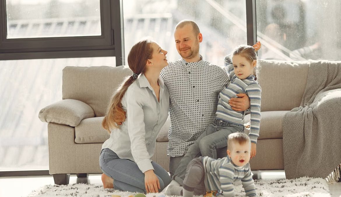 Cheerful mother and father smiling at each other while little cute boy crawling on floor and preschool girl in striped turtleneck raising arm and looking at camera