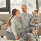 Cheerful mother and father smiling at each other while little cute boy crawling on floor and preschool girl in striped turtleneck raising arm and looking at camera