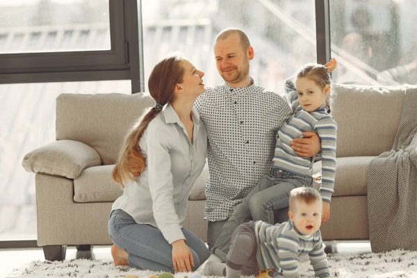 Cheerful mother and father smiling at each other while little cute boy crawling on floor and preschool girl in striped turtleneck raising arm and looking at camera