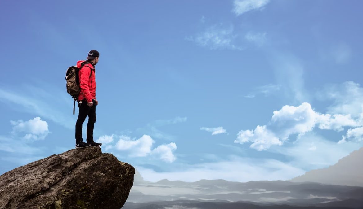 Hiker in red jacket stands on a cliff edge, overlooking a scenic mountain view under a bright blue sky.