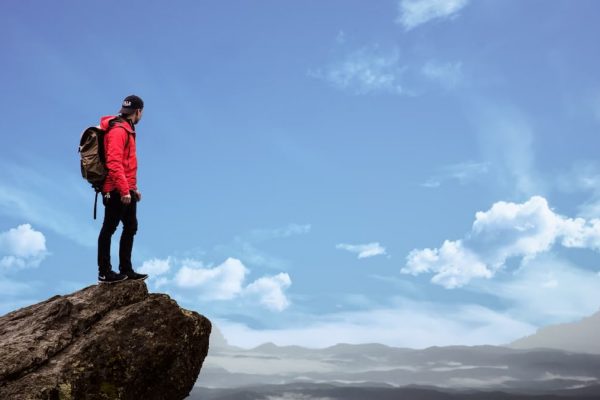 Hiker in red jacket stands on a cliff edge, overlooking a scenic mountain view under a bright blue sky.